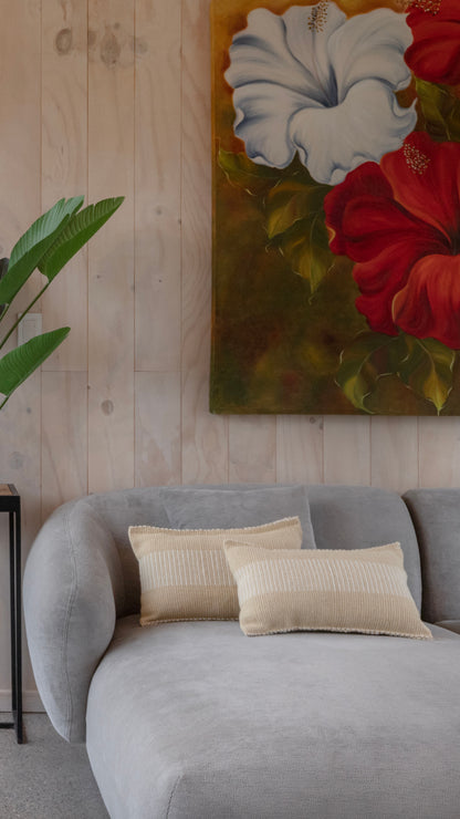 A modern gray fabric sofa in a living room setting, styled with two beige textured throw pillows. Above the sofa is a large vertical canvas painting depicting white and red hibiscus flowers on a green and gold background, set against a light wood-paneled wall.